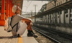 Viajante com mochila sentado na plataforma de uma estação ferroviária aguardando o embarque, ilustrando como viajar de trem na Itália com tranquilidade.